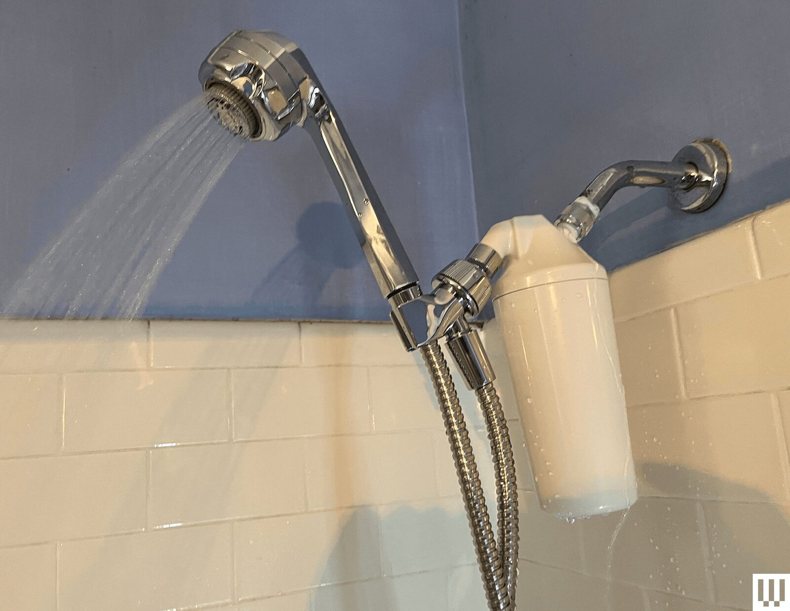 Silver elongated showerhead turned on with white tiles in the background and a white filter attachment connected to the...