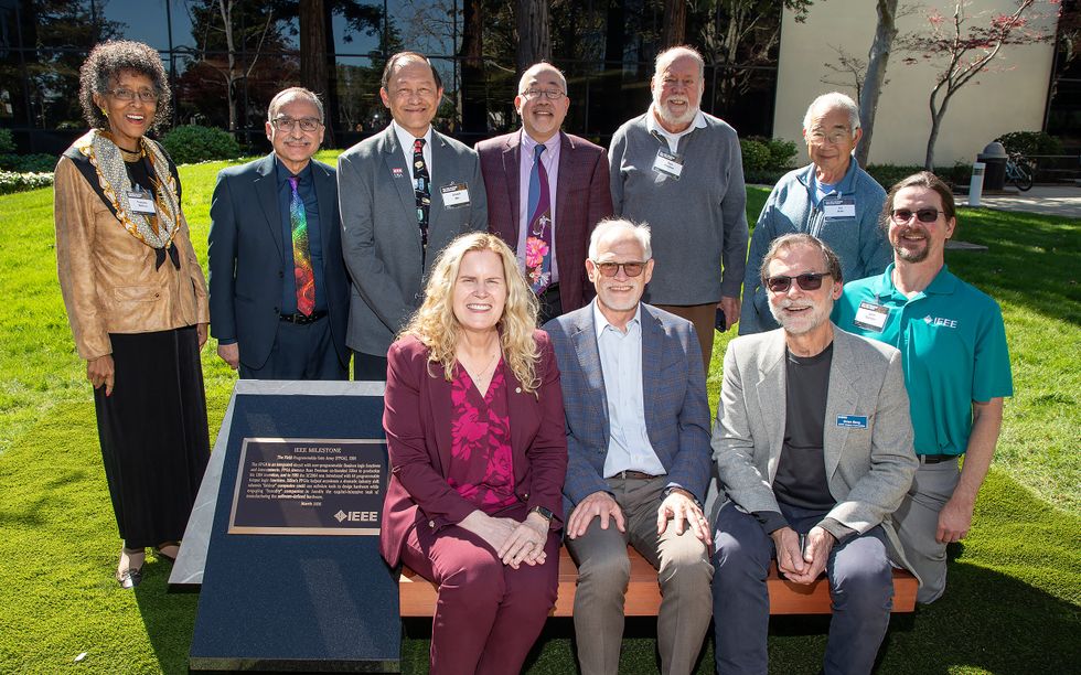 A group dressed in business casual attire smiling and posing together around an outdoor bench adorned with a plaque.