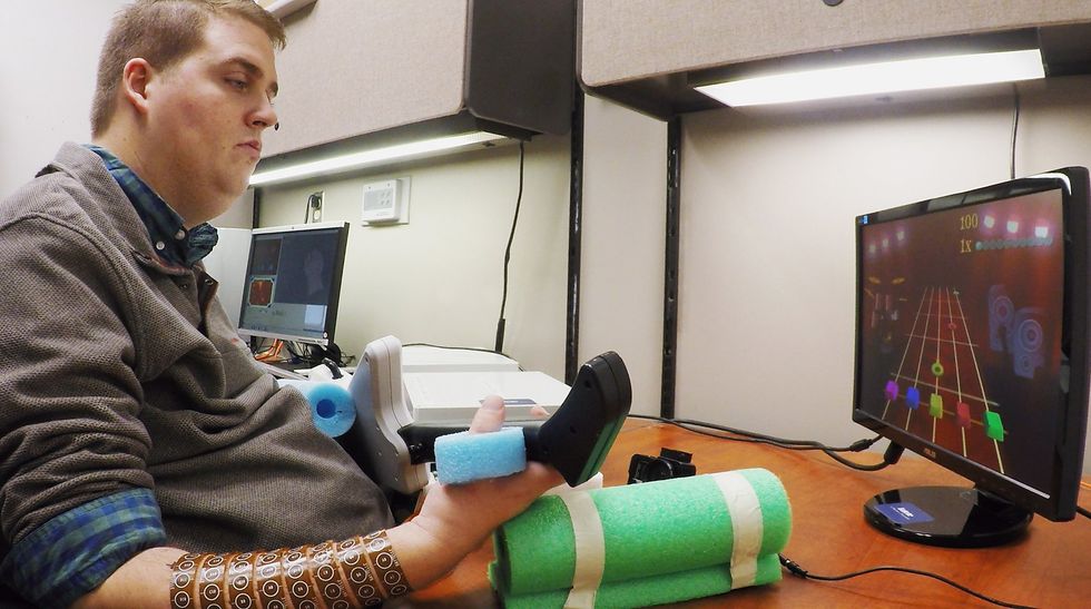 A man seated at a desk has electronics wrapped around his right arm. Heu2019s holding a device shaped like a guitar and looking at a screen showing the fretboard of a guitar.
