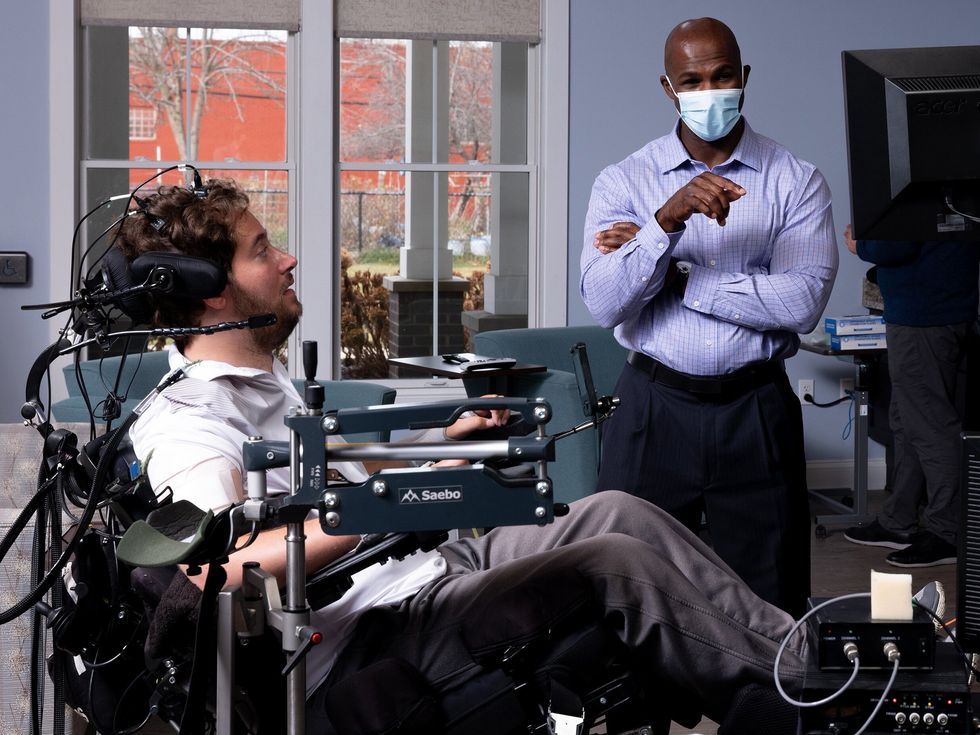 A man sits in a wheelchair surrounded by screens and electrical equipment. A device is attached to the top of his head, and a wire extends from it. Two other men stand in the room wearing masks.