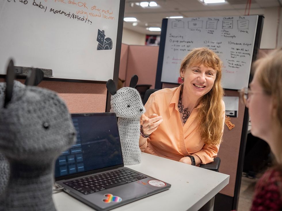 A smiling blonde woman gestures at a customizable tabletop robot that wears a knit outfit of a cute animal over its shell.