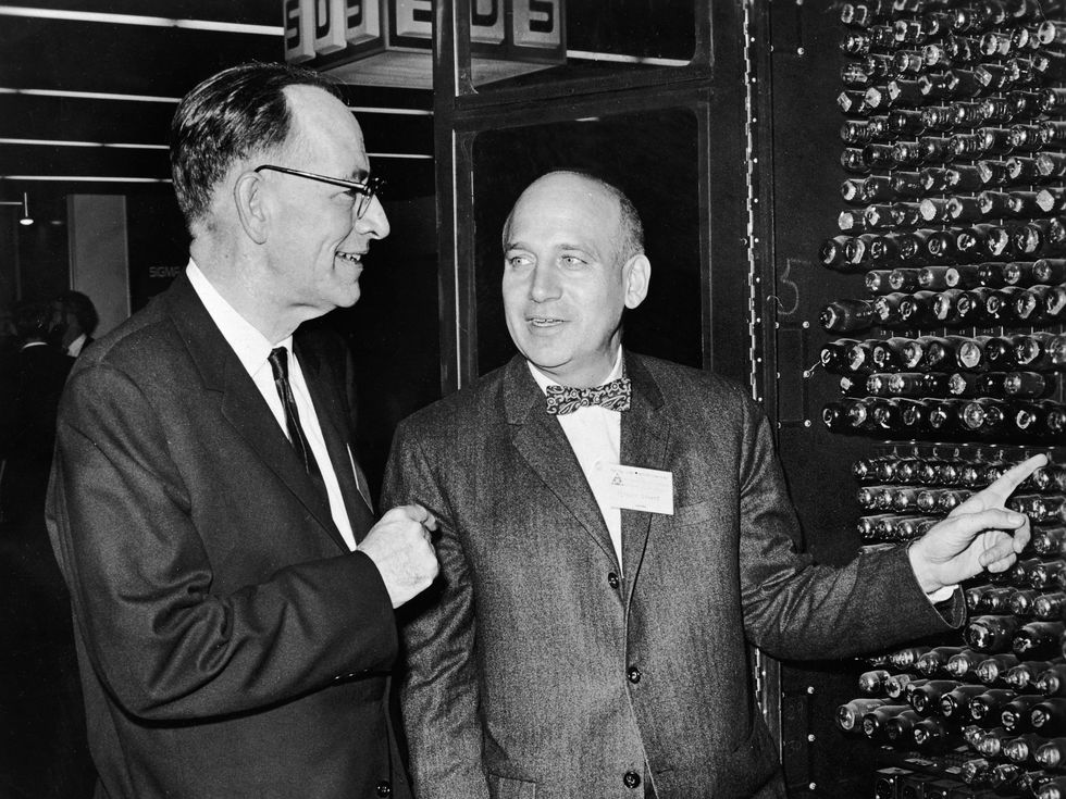 Black and white 1960s image of two white men in suits looking at a wall of computer controls.