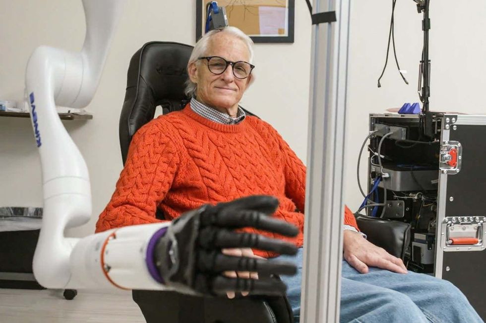 Elderly person in orange sweater sits as robotic arm with black hand extends forward