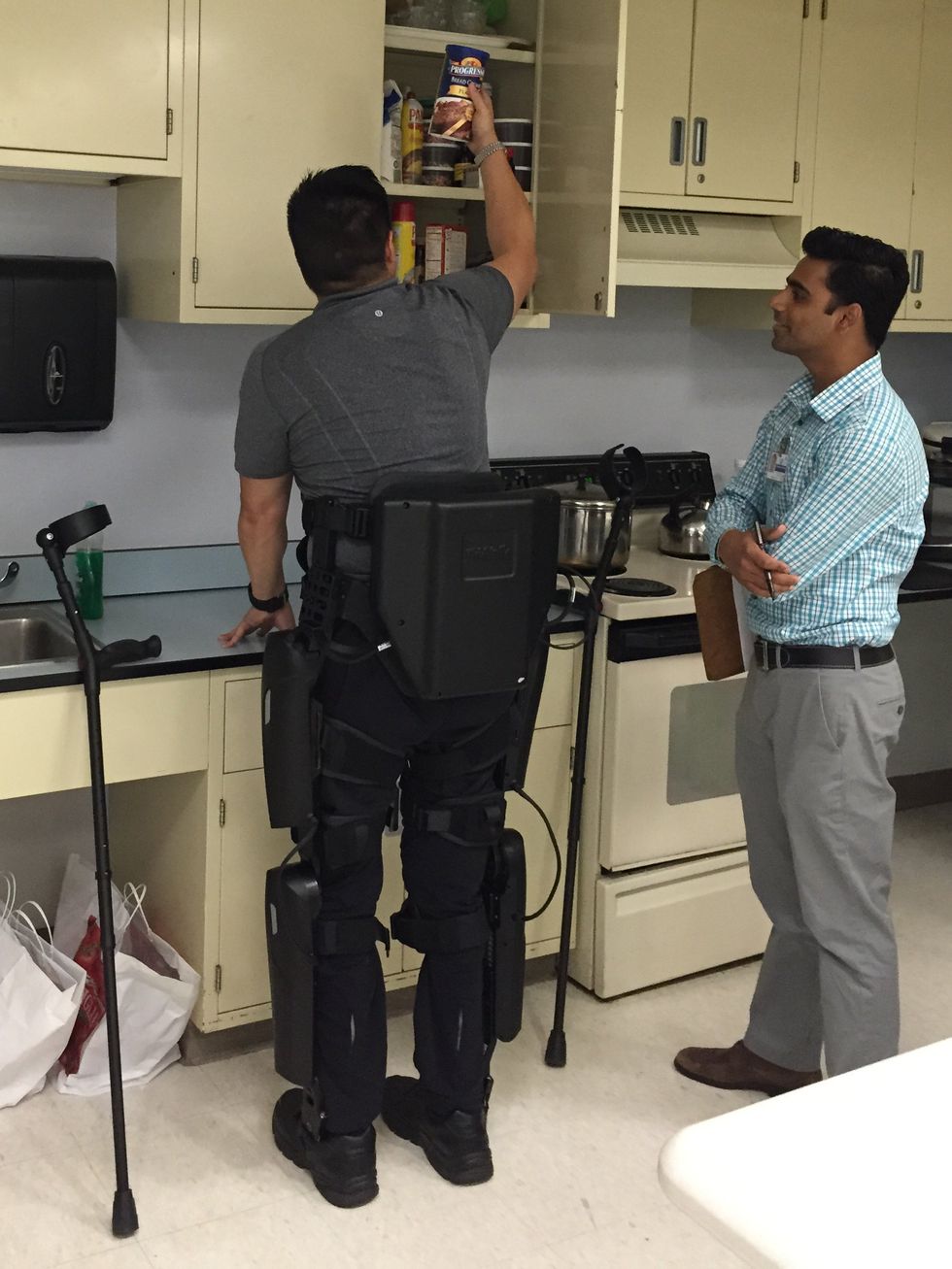 Man in a leg exoskeleton reaches into a kitchen cabinet while another observes.