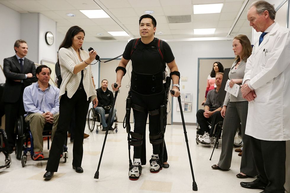 Man in robotic exoskeleton walks with canes during rehab demo as clinicians observe