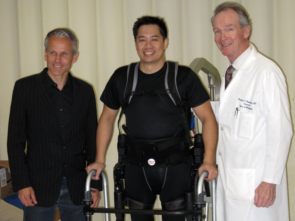 Patient in exoskeleton uses walker, flanked by doctor in lab coat and man in suit