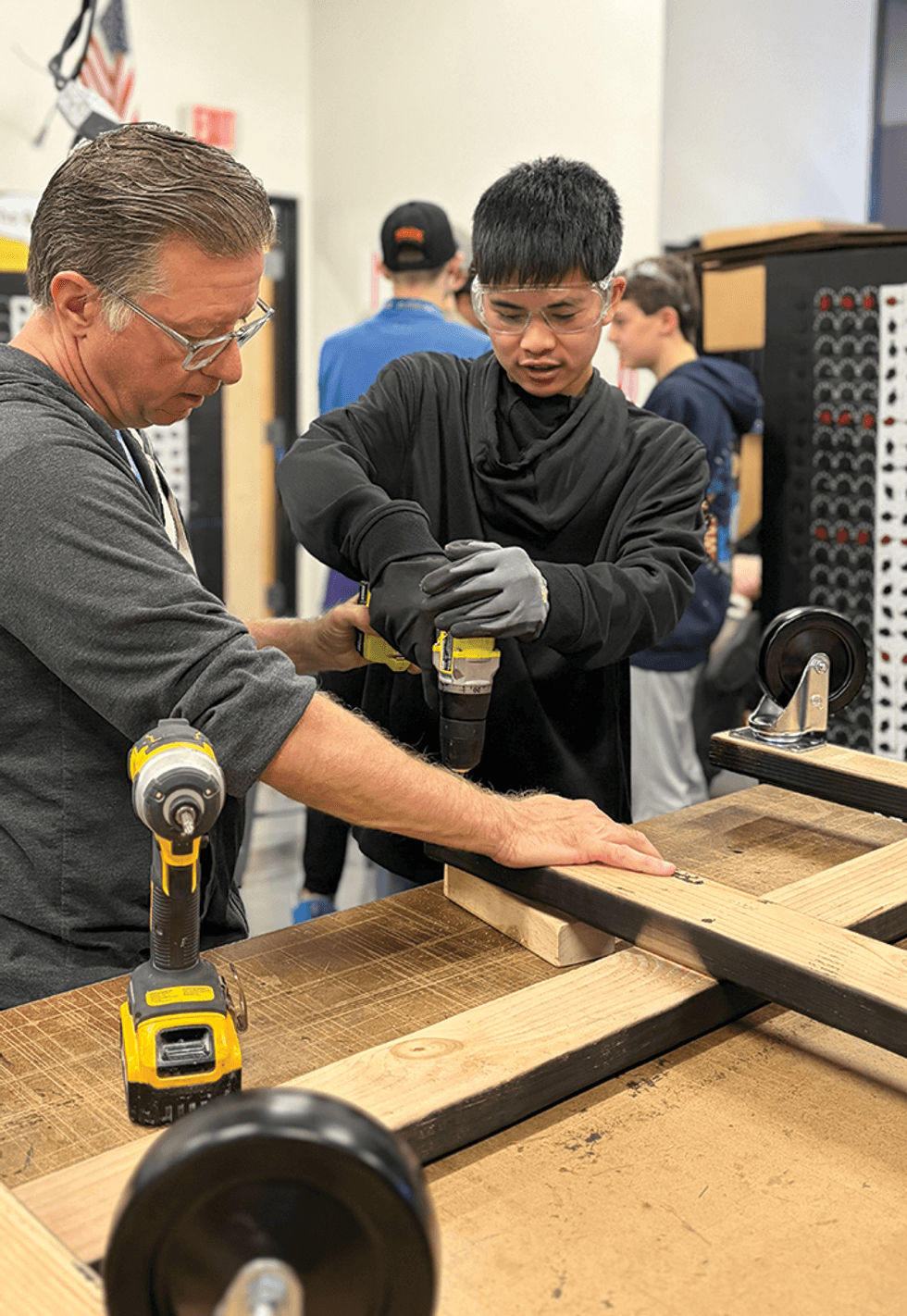 Student using power drill on wood under instructoru2019s guidance in workshop.