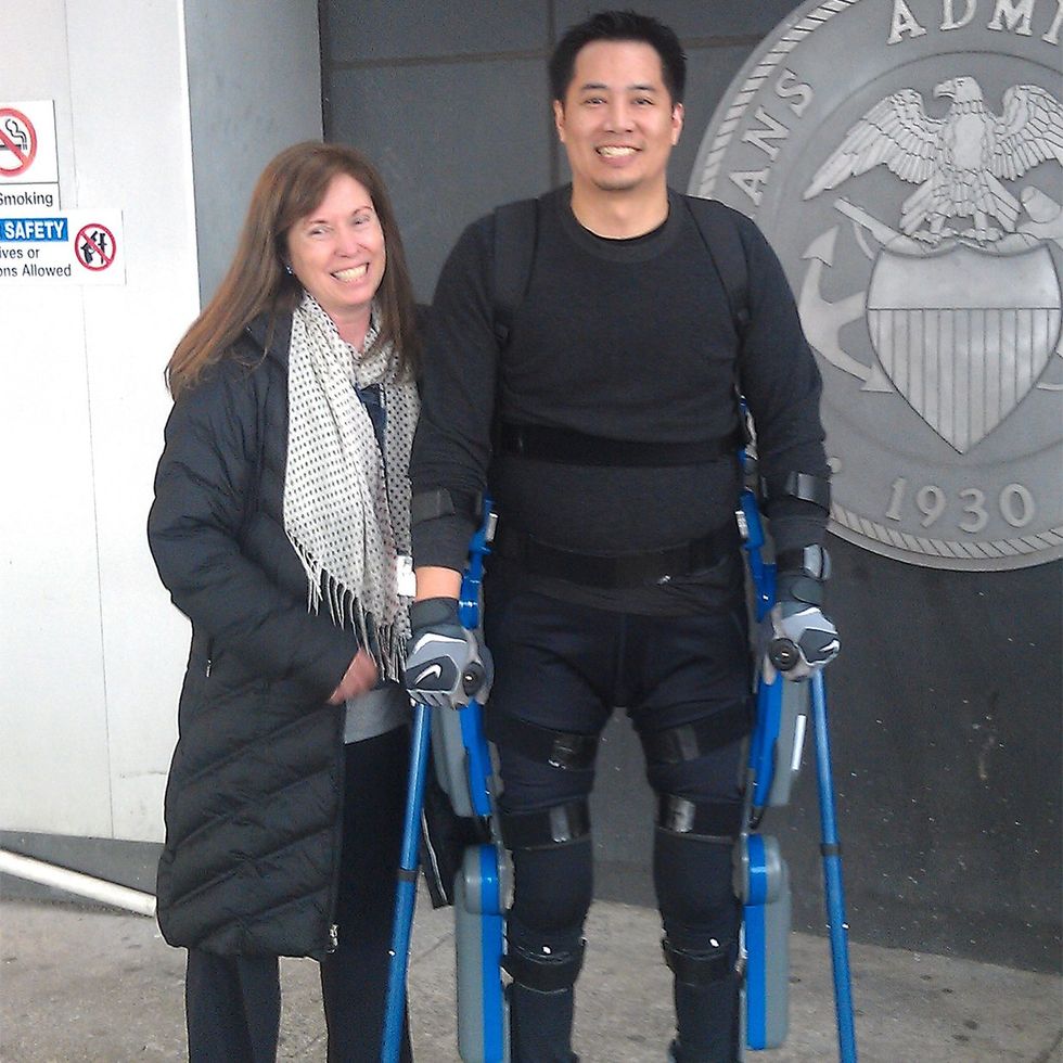 Two people stand outside; one uses blue exoskeleton crutches for mobility.