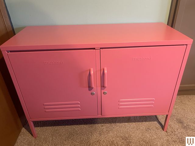 Mustard Made Lowdown Locker, a pink metal locker with two front doors, up against a wall while sitting on beige carpet